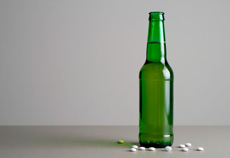 Зелёная стеклянная бутылка пива стоит на серой поверхности среди рассыпанных белых таблеток
